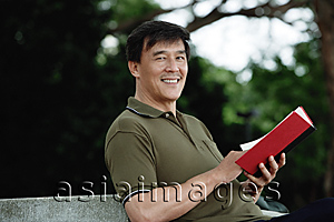 Asia Images Group - Man sitting on bench in park, reading, looking at camera smiling