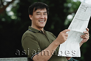 Asia Images Group - Man sitting on bench in park reading newspaper, smiling at camera
