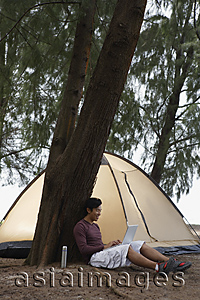 Asia Images Group - Man leaning against tree working on laptop, camping