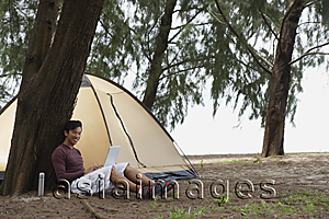 Asia Images Group - Man leaning against tree holding laptop, camping on beach