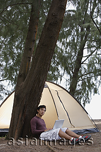 Asia Images Group - Man leaning against tree holding laptop, tent in background