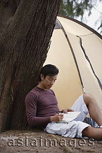 Asia Images Group - Man leaning against tree writing in journal, notebook, with tent in background, camping