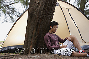 Asia Images Group - Man leaning against tree listening to music on MP3 player, camping with tent