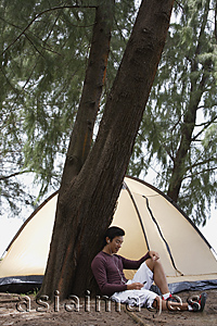 Asia Images Group - Man leaning against tree listening to music on MP3 player, tent in background, camping