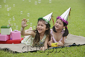 AsiaPix - Two girls wearing party hats lying on picnic blanket, looking at bubbles