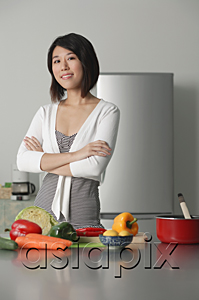 AsiaPix - Young woman in kitchen, arms crossed, vegetables on the table in front of her