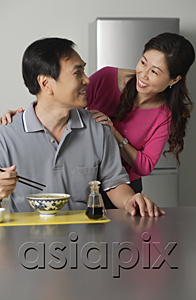 AsiaPix - Mature man in kitchen, eating a bowl of noodles, turning to look at woman standing behind him