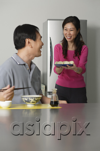 AsiaPix - Mature couple in kitchen, man sitting in front of a bowl of noodles, woman behind him holding a plate of food