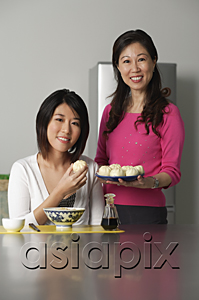 AsiaPix - Mother and daughter in kitchen, younger woman sitting, older woman standing beside her holding a plate of Chinese buns
