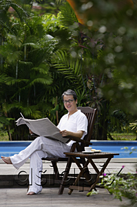 AsiaPix - Mature man sitting by swimming pool, reading newspaper