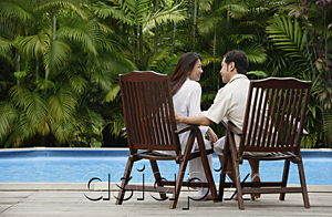 AsiaPix - Couple sitting by swimming pool,  looking at each other, rear view