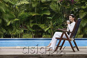 AsiaPix - Woman sitting by swimming pool, drinking from cup, looking away