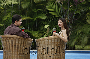 AsiaPix - Couple sitting by swimming pool in rattan chairs, woman turning to look at camera