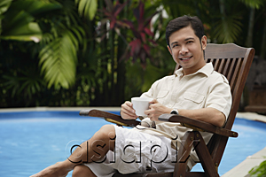 AsiaPix - Man sitting by swimming pool, holding a cup of coffee