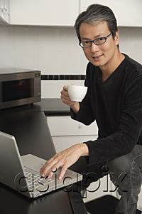 AsiaPix - Man in kitchen with laptop / computer, looking at camera, sitting on stool, smiling and holding coffee cup.