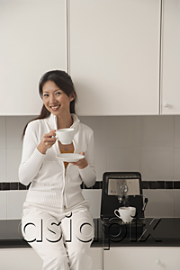 AsiaPix - Woman sitting on counter in kitchen next to cappuccino machine, holding coffee, smiling, looking at camera.