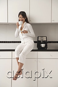AsiaPix - Woman sitting on counter in kitchen next to cappuccino machine, drinking coffee and looking at camera.