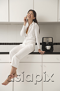AsiaPix - Woman sitting on counter in kitchen next to a cappuccino / coffee machine, talking on phone and smiling
