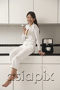 AsiaPix - Woman sitting on counter in kitchen next to a cappuccino / coffee machine, sending / receiving text message