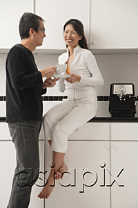 AsiaPix - Woman sitting on counter in kitchen with man standing next to her, both drinking coffee, looking at each other.