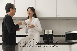 AsiaPix - Woman sitting on counter in kitchen with man standing next to her, looking at each other, holding coffee cups and smiling