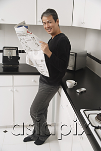 AsiaPix - Man in kitchen, leaning on counter, holding newspaper and looking at camera smiling.