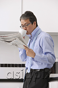 AsiaPix - Man in kitchen getting ready for work, drinking coffee and reading newspaper, tie loose around neck