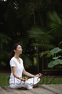 AsiaPix - Woman in tropical setting, meditating on porch, eyes closed, in yoga OM posture, side view.