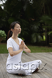 AsiaPix - Woman sitting on porch, in tropical setting.  legs crossed, hands in namaste, yoga posture. eyes closed