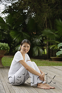 AsiaPix - Woman sitting on porch in tropical setting, arms wrapped around legs, looking at camera, smiling