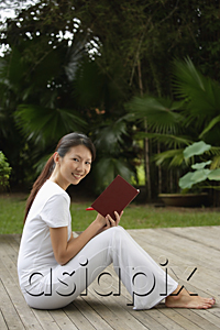 AsiaPix - side profile of woman sitting on porch in tropical setting, writing in journal, diary, and looking at camera, smiling
