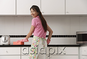 AsiaPix - woman standing in kitchen, wearing gloves and apron and cleaning counter with big pink sponge, looking over shoulder at camera, smiling.