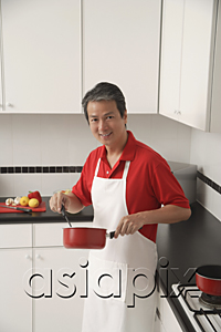 AsiaPix - Man in kitchen cooking with sauce pan, looking at camera, wearing apron