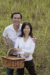 AsiaPix - Man and woman standing outdoors in nature, holding picnic basket, smiling at camera
