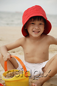 AsiaPix - Young boy sitting on beach, wearing read hat and holding yellow beach bucket