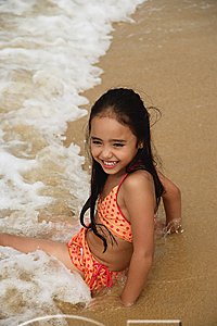 AsiaPix - Young girl sitting on beach with waves crashing over her, smiling
