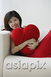 AsiaPix - Young woman sitting on sofa with heart-shaped cushion