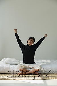 AsiaPix - Young man sitting on bed, stretching arms