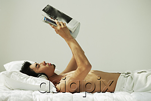 AsiaPix - Young man lying in bed, reading and listening to music