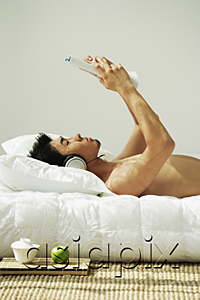AsiaPix - Young man lying in bed, reading and listening to music