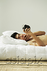 AsiaPix - Young man lying in bed, yawning and holding alarm clock
